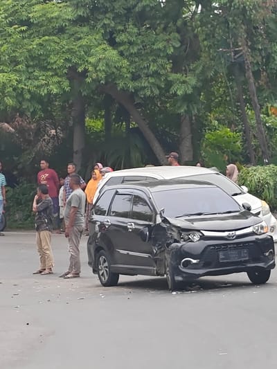 On a paved road in Meunasahgarot, Indonesia, a black car with significant front-end damage is parked. The damage appears to be the result of a collision. A white car is parked behind it, and a group of people, including men, women, and children, are standing on the side of the road, observing the scene. The environment is lush with trees and foliage lining the background. The road surface is asphalt and appears to have scattered debris.