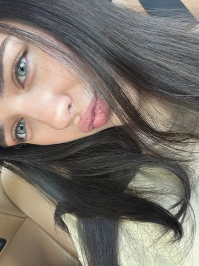 A close-up, upside-down, or sideways selfie shows a person's face, primarily focusing on their eye and mouth, with their dark hair obscuring parts of their face and framing the shot. The person has light skin and light green eyes, with dark eyebrows and subtle makeup. Their lips are slightly parted, showing a pinkish hue. The background appears to be the textured, light brown leather interior of a car seat, suggesting the photo was taken inside a vehicle. The lighting indicates it is daytime, possibly outdoors, as there are highlights and shadows on the hair and face. The overall mood is casual and personal, characteristic of a selfie. No visible text is present in the image. The location context is the City of Leduc, Canada.