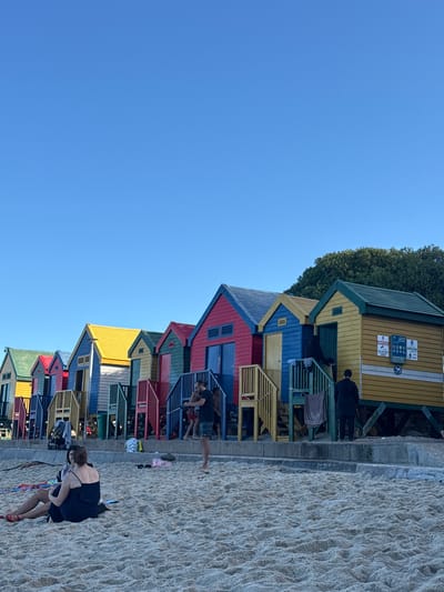 The image captures a row of vividly painted beach huts at Muizenberg, South Africa, situated alongside a sandy beach under a clear blue sky. The huts are colored in red, yellow, blue, green, and other bright hues. People are walking along the beach next to the huts. A woman in a dark dress stands on the sand. Several other people appear near the huts and along the walkway. The photograph is taken at ground level, with the huts and beach dominating the lower half of the frame and the expansive blue sky filling the upper half.
