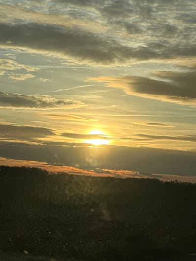 The image displays a serene landscape captured during either sunrise or sunset, characterized by a dramatic sky and a prominent sun. The original image is oriented vertically, but viewing it rotated 90 degrees counter-clockwise reveals the true horizontal composition.

In the upper portion of the correctly oriented image, a vast sky dominates the scene. It is filled with various clouds, ranging from thin, wispy cirrus-like streaks to more substantial, textured formations. These clouds are illuminated with warm, golden, orange, and pale yellow hues, especially those closer to the sun. Further away from the sun, the clouds transition into lighter grey and soft blue tones, indicating the varying light absorption and reflection. Some faint, linear contrails are also visible against the sky, particularly in the mid-left area.

The sun itself is a brilliant, intense light source positioned slightly to the right of the center, close to the horizon. Its bright, almost blown-out white and yellow light casts a strong glow, creating a beautiful lens flare effect and bathing the surrounding clouds in radiant warmth. The light from the sun appears to be streaming through gaps in the clouds, creating visible shafts of light.

Below the illuminated sky and sun, a clear horizon line separates the sky from the land. The land appears as a dark, silhouetted band, suggesting a distant treeline or a low-lying landscape, possibly a forest or fields. The very top edge of this dark landmass catches some of the sun's warm light, exhibiting subtle reddish-orange tinges. The bottom third of the image is comprised of this dark land, which is largely devoid of detail due to being in shadow against the bright backlight of the sun. There may be some faint textural elements suggesting ground or dense foliage, but it remains predominantly dark.

The overall impression is one of natural beauty and tranquility, showcasing the vibrant colors and dynamic light play characteristic of a sun near the horizon. There are no people or specific activities visible, focusing solely on the natural elements of the landscape and sky. No text is visible in the image.