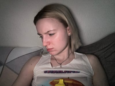 A person is lying down on what appears to be a bed or sofa. They are wearing a white t-shirt with writing on it and a necklace. The person has blond hair. The location is not discernible from the image, and there is no indication of Borisova Griva Massiv, Russia.