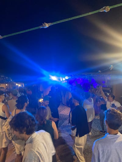 The image depicts a nighttime scene at a gathering or event in Agafay, Morocco. A crowd of approximately 15-20 individuals is visible, many of whom appear to be dancing or socializing. The lighting is predominantly blue and white, emanating from stage lights or spotlights, creating a vibrant and energetic atmosphere. Some individuals are dressed in casual attire, including shirts, shorts, and dresses. The background suggests an outdoor setting, possibly a desert camp or a venue with a rustic, open-air structure. A green cable is visible strung across the top of the frame, with small objects attached to it. The overall impression is one of a lively social event taking place after dark.