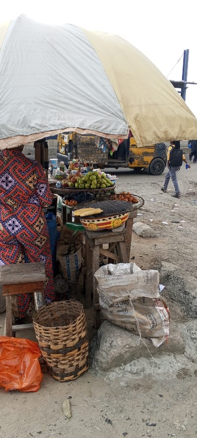 The scene portrays a roadside market stall in Awjaw, Nigeria, under an expansive, two-toned light grey and beige canopy. On the immediate left, a person is partially visible, wearing a vibrant garment with red, orange, and blue geometric and floral patterns.
The stall consists of wooden structures displaying various items. On an upper tier, a large basket holds numerous green apples or similar fruit, adjacent to a smaller quantity of red apples in another container. Below these, a decorative bowl with red, yellow, and orange markings features a black metal grill, upon which two pieces of yellow corn or plantain rest. Adjacent to this grill, a small, dark basket contains multiple small, round, reddish-brown items. Further back on the stall, assorted packaged goods and small containers are arranged on shelves.
At ground level, a large blue bag labeled "CEMENT" is visible, along with a white woven sack. An empty, woven wicker basket stands near an orange plastic bag. The ground is unpaved, consisting of dirt and gravel, with several large concrete blocks or rocks scattered throughout.
In the mid-ground, a heavily used yellow commercial vehicle, possibly a small bus or tricycle taxi, is positioned on a paved road. In the background, a person wearing a yellow shirt, grey pants, and a black backpack walks from right to left. The sky overhead appears overcast.