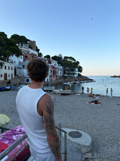 The image depicts a scenic view of the Aiguablava cove, located in the municipality of Begur, Spain. In the foreground, a young man with tattoos on his arm is seen from the back, leaning against a railing and looking out at the cove. He is wearing a white tank top and striped shorts. In front of him are parts of a purple kayak and a yellow flotation device.
The cove itself is characterized by its pebble beach, where several people are scattered, some sunbathing, others standing or walking. A collection of small boats are moored in the calm, blue water. Behind the beach, a hillside is covered with dense green trees and dotted with whitewashed houses with colorful accents, characteristic of the Mediterranean architectural style. A small, ancient-looking tower is visible on the hill, adding to the historical charm of the location. The sky is clear and blue, with a bird in flight visible in the distance. The overall atmosphere is serene and idyllic, capturing a typical summer day in this picturesque coastal area.
