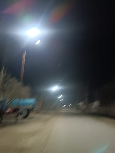 A blurred nighttime street view features evenly spaced streetlights illuminating a dirt road. Trees are visible on the left, partially obscuring a blue trailer-like structure. The sky is dark and clear. The photograph's blur suggests movement or a fast shutter speed. The scene creates a sense of rural isolation with the stark contrast of bright lights against the darkness.