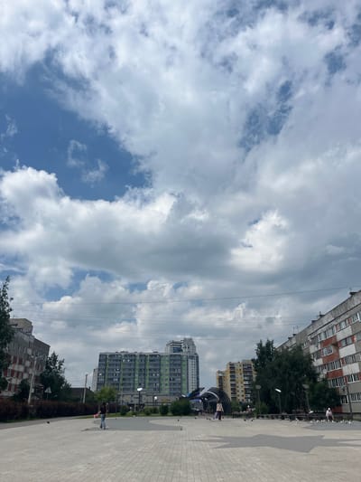 In the city of Imeni Sverdlova, Russia, a public plaza is depicted on a cloudy day. A large, modern, multi-story apartment building with a green and gray facade dominates the central background. To its left, a older, gray apartment building with red-accented windows is visible, partially obscured by trees. To the right, a weathered, red and gray apartment building with numerous windows is prominent. Several power lines crisscross the sky, stretching horizontally across the frame. The plaza itself is paved with light-colored interlocking stones, forming a large, star-shaped pattern in the foreground. A few scattered individuals are present in the plaza, including one person walking away from the camera with a pink object, and another person sitting on a bench. Birds are scattered across the plaza, some in flight and others on the ground. Trees and bushes line the edge of the plaza, separating it from the surrounding buildings.
