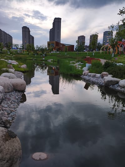 The image depicts a modern park in Jiguan District, China, at dusk. The foreground is dominated by a calm body of water with reflections of the surrounding environment. The water's edge is lined with large rocks and smaller pebbles, with patches of tall green grass growing between them. In the middle ground, a lush green lawn is dotted with trees and decorative elements, including colorful arched structures on the right. Beyond the park, several tall, modern residential buildings are visible against a cloudy sky. A low-rise building with a modern architectural design is situated among the trees. Several figures, appearing as small silhouettes, are scattered throughout the park, suggesting it is a public space. The overall scene conveys a sense of urban development and leisure.
