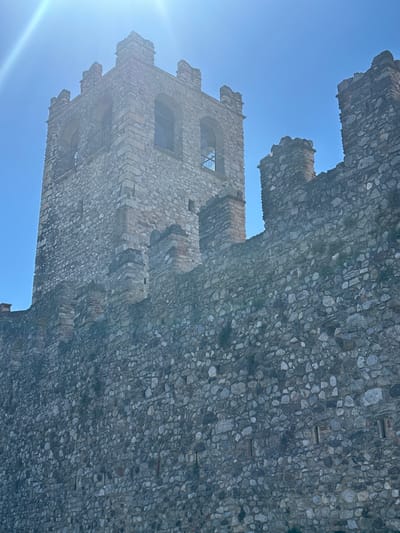 The image depicts the exterior of a stone castle or fortress, specifically the Torre Civica (Civic Tower) and its surrounding walls in Desenzano del Garda, Italy. The sky is a clear, bright blue, with a strong sun creating a lens flare effect in the upper left quadrant of the image. The tower, constructed of rough-hewn stones of varying sizes and shades of grey and brown, has crenellated battlements at its top. Two arched openings are visible in the upper section of the tower, hinting at windows or access points. The defensive wall in the foreground, also made of stone, features crenellations and appears to be quite substantial. Small patches of greenery are visible growing from the stone in various places on the wall. The overall impression is one of historical architecture bathed in bright sunlight. There are no people or animals visible in the image.
