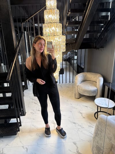 A woman stands on a marble-tiled floor, taking a mirror selfie. She is wearing black leggings, black Crocs, and a black top with a black cardigan draped over her shoulders. She has long brown hair and is wearing earrings and a necklace. In the background, there is a black metal staircase with diamond plate steps, a tall, multi-tiered crystal chandelier, and a light gray armchair next to a small circular table with a glass of water. The location is stated to be in the village of Ukromne, Simferopol Raion, Crimea.