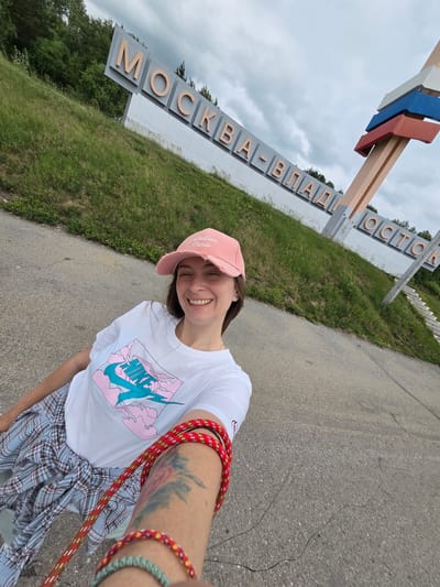A young woman with a pink baseball cap and a white t-shirt with a Nike logo and a pink cloud design is smiling at the camera. She is wearing a plaid shirt tied around her waist. A red rope is wrapped around her arm, and she has a tattoo on her arm. The background features a large sign that reads "МОСКВА-ВЛАДИВОСТОК" (Moscow-Vladivostok) in large letters. The sign is decorated with the Russian flag colors. The location is the city of Nikolaevka, Russia. The environment is outdoors with a cloudy sky and green grass.