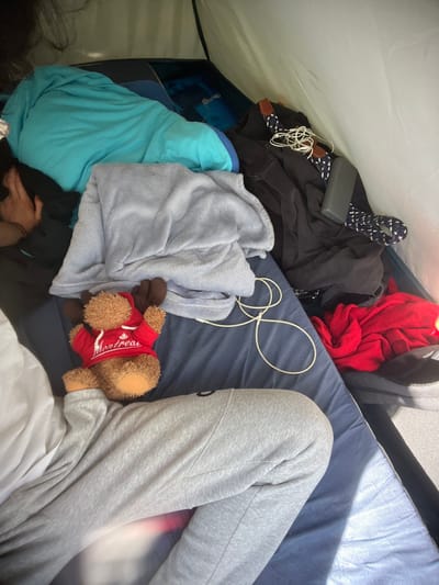 The image displays the interior of a tent, located in Sint-Pieters-Leeuw, Belgium. The scene captures a close-up, angled view looking down into the tent's living space.
On the left side of the frame, the lower body of an individual is visible, dressed in light grey sweatpants, lying on a dark blue sleeping mat. A hand with a bracelet is partially visible in the upper left corner, obscured by a light blue sleeping bag or large blanket. A light grey fleece blanket is folded and placed centrally over the sleeping mat.
Positioned on the dark blue sleeping mat, adjacent to the visible person's legs, is a brown stuffed moose wearing a red hoodie with "Montréal" text on it. White wired earphones are loosely coiled on the sleeping mat to the right of the stuffed animal.
The right side of the tent is occupied by a collection of personal items, including various dark-colored garments, a crumpled red item of clothing, and a dark grey power bank. A patterned dark blue and white strap is also visible among these items. The tent's light-colored fabric walls are visible on the right and top, illuminated by natural light entering from the right.