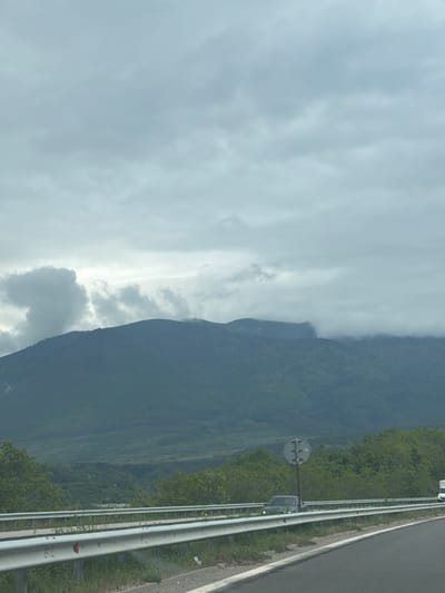 The image shows a mountainous landscape with a roadside scene. The sky is overcast with gray clouds. A dark, tree-covered mountain rises on the right side of the frame, its peak obscured by clouds. A highway runs parallel to the mountain, with a metal guardrail visible along its edge. A circular road sign is present beside the road. The location is likely near Djerman, Bulgaria, considering the mountain and roadside infrastructure. No people are visible.