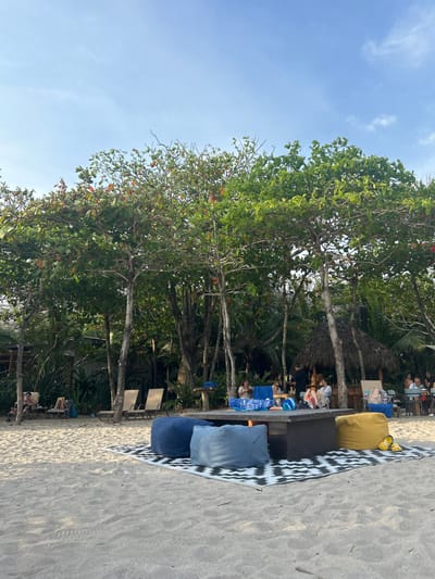 The scene is a beachside picnic area. There are several people gathered around a large wooden table covered in a black and white tablecloth. There are a few blue cushions scattered on the ground around the table, and one yellow cushion. In the background, there are palm trees and some people sitting at a bar. The beach is white sand and the sky is blue. The scene is located in Santa Teresa, Costa Rica, specifically at the coordinates latitude: 9.635, longitude: -85.161.