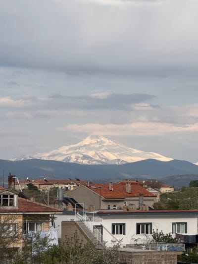 The image captures a view from Ürgüp, Turkey, showcasing a blend of urban development and natural landscapes. A snow-capped mountain serves as a backdrop to the town. To the right, a multi-story white building with dark-framed windows and a white balcony stands tall, adjacent to rooftops covered with red tiles. The building appears inhabited. The surrounding environment also reveals other buildings, varying in color and design. The sky above is partially cloudy, adding depth to the scenery.