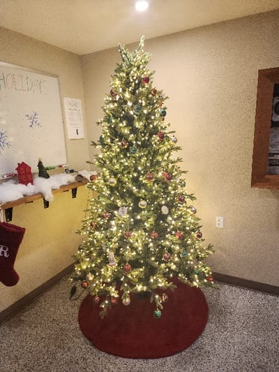 A well-lit Christmas tree adorned with colorful ornaments and white lights sits in a room with a red tree skirt. A whiteboard with holiday-themed drawings and the word "HOLIDAYS" is nearby, along with Christmas decorations on a wooden shelf. The room features a patterned carpet and beige walls. A framed object is mounted high on the wall near the ceiling.
