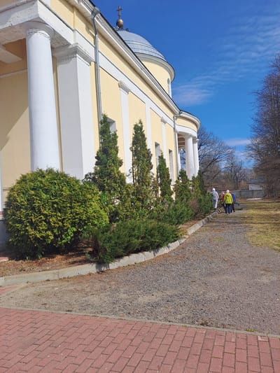 The image shows an exterior view of a building with classical architectural elements, including columns, a dome, and a cross on top. The building is pale yellow with white trim. In front of the building is a row of bushes. A path made of gravel runs parallel to the bushes. A red brick walkway is to the right of the gravel path. In the background, there are a few people and bare trees. The sky is blue with wispy clouds. This structure is located in the city of Ivashevo, Russia.