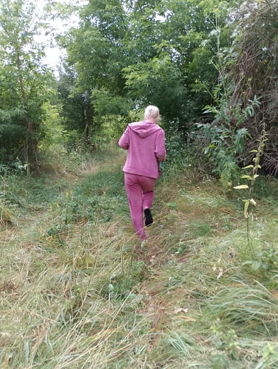 A woman with blonde hair is walking away from the camera into a grassy, wooded area. She is wearing a pink tracksuit and black shoes. The woman is walking on a path through tall grass and trees. The scene appears to be a rural area, possibly a park or a forest. The sun is shining, indicating it is daytime. There are no visible text or signs in the image. The overall mood is peaceful and natural.