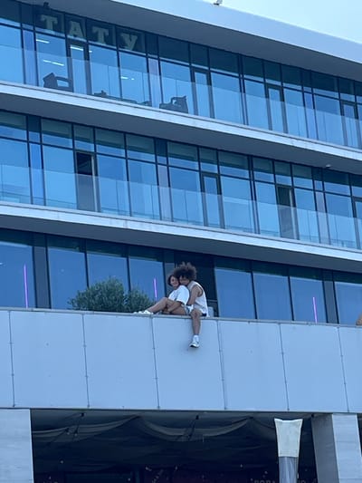 A medium shot shows two individuals sitting on the ledge of a modern building, facing left. The building has multiple floors with large, reflective blue windows. The word "TATY" is visible in large green letters above the windows on the top floor. A small bush is located behind the couple. The individual on the left has dark, curly hair and is wearing a white tank top and white shorts. They are leaning their head on the shoulder of the other individual. The individual on the right is wearing a white tank top and white shorts with blue trim. They are sitting with their legs dangling over the edge. The overall environment appears to be an urban setting, possibly Ol'ginskii, Russia, given the building's architecture and the presence of other structures below. The scene captures a moment of intimacy and relaxation.
