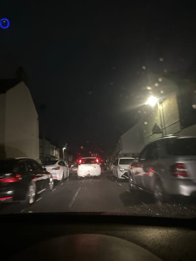 The image depicts a nighttime street scene in what appears to be a residential or mixed-use urban area of Gillingham, United Kingdom, viewed from the interior of a vehicle through a wet or dirty windshield. Traffic is present and appears to be stationary or moving slowly.
In the foreground, a dark dashboard or steering wheel is visible at the bottom of the frame. The road surface shows reflections consistent with moisture or rain, and scattered debris is visible.
Multiple vehicles are present. On the immediate left, the rear of a dark-colored sedan is partially visible. Further ahead on the left, a white sedan is seen. In the center, a white compact car, possibly a Fiat 500, faces away from the camera, with its red brake lights illuminated and a visible tow hitch. Light-colored marks or debris are present on its rear bumper and boot lid. On the immediate right, a light-colored (possibly silver) sedan is visible with motion blur, suggesting movement. Its rear taillights are active. Numerous other vehicles are indicated by distant red taillights further down the street.
The street is lined with multi-story buildings on both sides. On the left, a light-colored building with a dark roofline is prominent. On the right, a darker building is partially obscured by a bright streetlight emitting significant glare and lens flares on the windshield. Other streetlights are visible further down the road, casting illumination on the street and buildings. The sky is dark and devoid of visible stars. A distinct blue, circular light anomaly is present in the upper left corner of the frame. No individuals are clearly visible. The precise street name within Gillingham is not identifiable from the provided visual information.