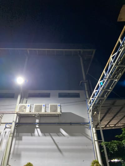 This is a medium-angle, low-light, full-shot photograph of an industrial building's exterior wall at night in Gambut, Indonesia. The wall is painted a light gray and features three white outdoor air conditioning units mounted on a silver rack. Above the air conditioners, there are three horizontal, rectangular windows. A bright, white security light illuminates the scene from the left. Various pipes, some white and some blue, run vertically and horizontally along the wall and the building's structure. To the right, a section of a light-colored metal walkway or canopy, supported by a truss structure, extends upwards and diagonally across the frame. The background is a dark night sky, with some faint atmospheric light visible near the security light and possibly reflecting off the metal structure. In the lower right corner, the top of a green leafy tree is visible. No people are present in the image.