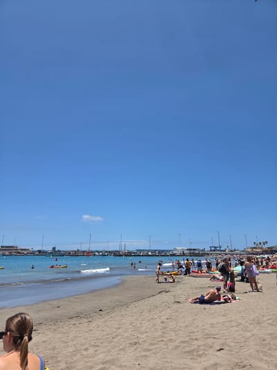 The image depicts a beach scene in Chayofita, Spain on a clear day. The sky is a bright, cloudless blue, taking up a significant portion of the frame. The beach is sandy and occupied by numerous people sunbathing, walking along the shore, and swimming in the sea. Along the edge of the beach, a series of lampposts are visible. The water appears calm with small waves. Some individuals are kayaking and there are a couple of people in the foreground.