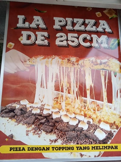 This is a promotional poster for a pizza, likely displayed at a food establishment in Kecamatan Panji, Indonesia. The image features a close-up of a long, rectangular pizza. A hand is visible on the left, pulling up a slice of the pizza, creating numerous long, stretchy strings of melted cheese.
The pizza is distinctly divided into two halves with different toppings. The half from which the cheese is being pulled appears to be a savory pizza, though specific toppings are obscured by the extreme cheese pull. The other half of the pizza is a dessert pizza, generously covered with chocolate sprinkles, dark crumbled cookies or brownies, and numerous small, toasted marshmallows.
The background of the poster is a reddish-brown color, adorned with scattered depictions of small cheese cubes and green chili peppers or herbs. Bold, white capital letters at the top of the poster read "LA PIZZA" and "DE 25CM," indicating the name of the pizza and its approximate length. A bright yellow banner at the bottom of the image contains black text that says "PIZZA DENGAN TOPPING YANG MELIMPAH," which translates to "PIZZA WITH ABUNDANT TOPPINGS." The overall image emphasizes the rich toppings and the appealing, stretchy quality of the cheese.