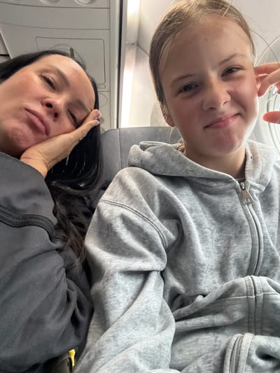 The image shows two individuals, likely a mother and daughter, seated on an airplane, as indicated by the airplane cabin interior visible in the background. The woman on the left appears tired, with her head resting on her hand and her eyes closed. She has long dark hair and is wearing a dark gray top. The girl on the right is looking at the camera with a slight smile, and her hand is near her face. She has blonde hair and is wearing a gray hooded sweatshirt. The lighting suggests an indoor environment, possibly during daytime given the light coming from the window. There is no identifiable information that places this image in Shul'gino, Russia.