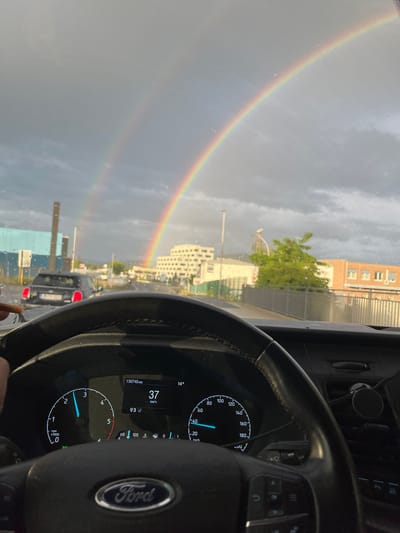 The image is a first-person perspective, taken from inside a Ford vehicle, looking through the windshield. The windshield is wet with raindrops. A double rainbow is visible in the sky. The road ahead is a multi-lane street with other vehicles. On the right side of the road, there are buildings. On the left side of the road, there is a tall industrial structure and a blue building. The steering wheel of the car is in the foreground, with the Ford logo clearly visible. The car's dashboard is also visible, showing the speedometer indicating 37 km/h and the tachometer. The overall scene suggests a drive through a city during or after a rain shower, with a beautiful natural phenomenon in the sky. The specific location is identified as the city of Aschaffenburg, Germany.