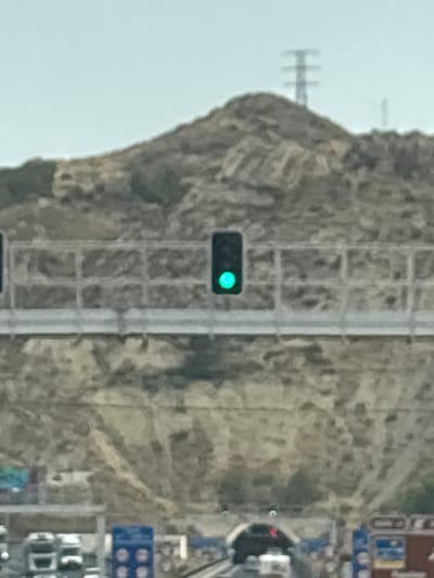 The image depicts a traffic light displaying a green signal, positioned horizontally on a light pole. This traffic light is situated on an elevated roadway with a metal railing. In the background, a rocky hill with sparse vegetation is visible, topped by a transmission tower. Below the elevated roadway, vehicles are seen on what appears to be a road or tunnel entrance. The overall scene suggests a highway or major road in a semi-arid or mountainous region. There are no people visible in the image. The location is identified as being in the city of Lorca, Spain.