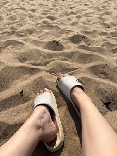 The image shows two feet wearing white slide sandals, partially submerged in rippled sand. The sand appears dry and sandy, with shadows indicating it is daytime. The perspective is a close-up shot, focusing on the feet and the texture of the sand. One foot is positioned higher in the frame, with the leg visible above the sandal. The other foot is lower down, with the heel slightly raised. The overall scene suggests a casual setting, likely a beach or a sandy area. There are no discernible emotions or specific events taking place, beyond the feet resting on the sand. The location context is Mikhalkino, Russia.