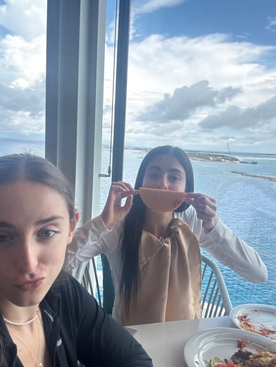 Two young adult females are seated at a white table in a dining area with large vertical windows overlooking the Berry Islands, Bahamas. The female on the left, partially visible in the foreground, has dark hair and blue eyes, wearing a black long-sleeved top, a white pearl choker, and a silver pendant necklace; her lips are slightly pursed. The female on the right, fully visible, has long dark hair, wears a white long-sleeved shirt, and a light beige or gold cloth bib. She holds a U-shaped taco shell or crisp to her mouth, mimicking a smile. On the white table are two white plates containing remnants of food, one with pasta and another with what appears to be taco or nacho ingredients. Through the windows, a cloudy sky is visible above a vast expanse of blue ocean. In the mid-distance, a narrow landmass with sparse vegetation and visible construction activity, including a large crane and barges, is present.