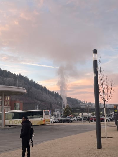 The image depicts an outdoor scene in Tuttlingen, Germany, at either sunrise or sunset, characterized by a pink and blue sky with scattered clouds and a visible aircraft contrail. A significant column of white vapor or smoke rises vertically from behind buildings in the mid-ground against a frost-covered evergreen hill in the background.

In the foreground, a dark-skinned male subject, wearing a dark puffer jacket, dark pants, and a dark backpack, stands with his back partially towards the camera, looking right. His right hand is raised, possibly holding a phone to his ear. To the right, a tall, dark grey cylindrical lamp post with an illuminated section stands beside a small, bare-branched tree.

The mid-ground features a white and yellow "move" branded bus parked or moving slowly to the left of the frame, near a building with a curved canopy and red brick facade. Behind the bus and extending to the right is a parking area filled with various vehicles, including cars and a white tanker trailer. A decorated Christmas tree with lights is visible within this parking area, in front of the hill. A blue sign with a white 'H' symbol, indicating a bus stop, is visible to the right. The background is dominated by a forested hill covered in a thin layer of white frost, likely indicating cold temperatures.