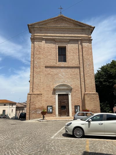 The image shows the facade of a brick church in the city of Barbara, Italy. The church is tall and narrow, with a triangular pediment at the top and a cross. The facade has a large wooden door and a single window above it. There is a potted plant with red flowers to the left of the door and a bulletin board to the right. In front of the church is a paved square with cobblestones. Several cars are parked in the square, including a white Alfa Romeo in the foreground. The sky is blue with some white clouds, and a few trees are visible to the right of the church. The overall impression is a sunny day in a quiet Italian town.