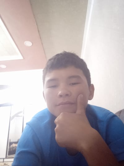 A young boy with dark hair, wearing a blue t-shirt, poses for a selfie. He has his right thumb placed under his chin. The background shows a ceiling with inset lights and a white wall. The setting appears to be indoors, possibly a home or room, located in Джапалак, Kyrgyzstan.