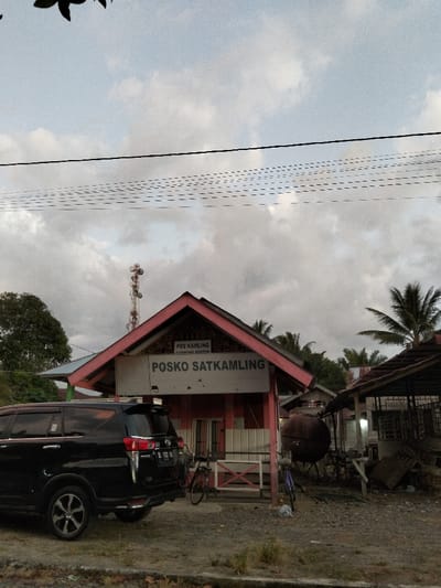 An outdoor scene in Panton, Indonesia, depicts a daytime view under an overcast sky. The central subject is a small, single-story building with a red-orange gabled roof and light-colored walls, identified by a sign that reads "POSKO SATKAMLING" and a smaller sign above it stating "POS KAMLING GAMBONG PANTON."
To the left of this building, a black Toyota Kijang Innova SUV (license plate BL 1722 NL) is parked, oriented with its rear facing the viewer. A red-framed bicycle is parked near the front of the "POSKO SATKAMLING" building. To the right of the building, a large, rusty, spherical tank is situated, with another bicycle, possibly blue, parked nearby. Further to the right, a rustic, open-sided structure with a dark roof is visible, surrounded by miscellaneous debris and materials.
In the background, a tall communication tower with multiple antennas rises behind the main building and to its left. Overhead, several power lines traverse the upper portion of the frame. Scattered palm trees and other foliage are visible behind the structures. The ground in the immediate foreground and around the buildings appears to be unpaved dirt or gravel. No individuals are visible in the scene.