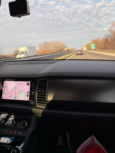 The image depicts the interior of a vehicle on a highway, viewed from the passenger's perspective looking forward through the windshield. The vehicle is located on a highway in Plovdiv Province, Bulgaria, specifically identifiable by "Plovdiv" and "с. Белащица" (Belashtitsa village) visible on the active navigation map, suggesting proximity to Trilistnik.
The vehicle's dashboard, dark gray with a carbon-fiber textured panel on the passenger side, occupies the lower portion of the frame. A central infotainment screen displays a navigation map with a pinkish background, indicating a speed of 142 km/h and a time of 7:54. Below the screen, climate control dials and buttons are visible. A black dash camera is mounted on the upper center of the windshield. In the lower left, charging cables and a white and blue packet (possibly wipes or tissues) are visible in a storage compartment. In the lower right, the passenger's right foot, wearing a dark shoe with white laces, is visible, along with a red object partially concealed within a light-colored, crumpled bag.
Outside the vehicle, a multi-lane highway stretches into the distance under a partly cloudy sky with patches of blue. Sunlight illuminates the scene, casting shadows on the dashboard. Leafless trees and sparse brown vegetation line both sides of the highway, separated by a metallic guardrail. On the left side of the highway, a white box truck is visible in the distance. Ahead on the highway, a dark-colored sedan travels in the left lane. Further down the road, a green highway sign with white lettering is visible on the right shoulder.