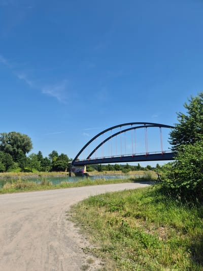 This image depicts a scenic view of a bridge arching over a canal, with a dirt path running alongside. The bridge, painted a dark color with red accents, features a striking curved design supported by numerous cables. Lush green trees and tall grass flank the path and canal, creating a vibrant natural setting. The sky above is a clear, brilliant blue, suggesting fair weather and a bright, sunny day, likely in the afternoon.

The image has been rotated 90 degrees counter-clockwise, so the bridge appears to span horizontally across the frame from left to right, with the canal running below it. The path is to the right of the canal. Graffiti is visible on the side of the bridge abutment, adding a splash of color to the scene. In the distance, two wind turbines can be seen peeking over the trees, hinting at a rural or semi-rural location. The overall impression is one of a peaceful, outdoor environment, possibly a recreational area for walking or cycling. No people are visible in the frame. The location is identified as Lüdinghausen, Germany.