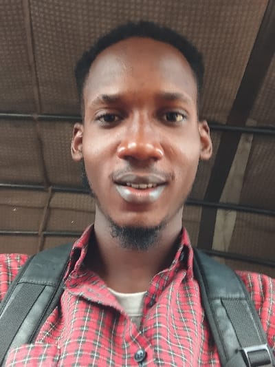 A young Black man wearing a red and black plaid shirt and black backpack smiles broadly. His skin is brown and slightly glossy. He has a short beard and black hair. The background appears to be a textured, neutral color with some linear elements. The angle of the image is slightly tilted.