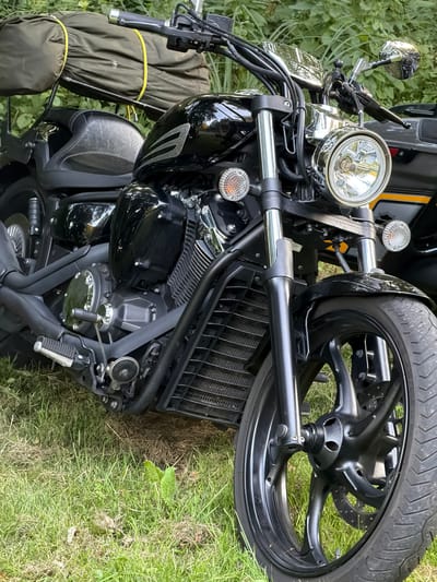 This media file is an overhead, angled shot of a black motorcycle parked on grass, likely in Mensdorf, Luxembourg. The motorcycle is the sole subject of the image, with its front wheel and part of the engine and handlebars visible. The lighting suggests it is daytime, with natural light illuminating the scene.
The motorcycle is black with chrome accents, including the handlebars, mirrors, and headlight casing. The front tire is large and appears to be in good condition. The engine is visible, showing intricate details of its construction. A black saddlebag is attached to the side of the motorcycle, secured with yellow straps. The motorcycle is positioned on a patch of green grass, with some areas of dry earth also visible. The background consists of out-of-focus greenery, likely trees or bushes, suggesting an outdoor setting. There are no people present in the image, and no specific activity or event is taking place; the motorcycle appears to be stationary. No visible text is present in the image. The overall impression is one of a parked motorcycle in a natural, possibly rural or park-like, environment.