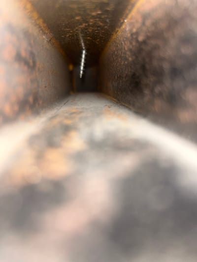 The image provides a close-up, internal perspective looking down a narrow, enclosed channel or pipe, likely made of metal. The interior surfaces of the channel exhibit significant texture, characterized by patterns of rust and grime in shades of dark brown, orange, and gray, suggesting an aged or industrial environment. The rough, granular quality of the walls is evident throughout.
Positioned horizontally and centrally deep within this confined space is a small, brightly reflective metallic object. This object clearly appears to be the threaded shaft of a screw or bolt, catching and reflecting light against the darker backdrop of the channel's interior.
The right side of the image is heavily blurred and out of focus, featuring broad strokes of gray, white, and soft orange hues. This blurred area suggests an external environment or a large object positioned immediately outside the channel's opening, contributing to the ambient light. No individuals or specific activities are depicted; the scene is static, focusing on the structural details of the channel and the small fastener within. There is no discernible text. While the precise nature of the structure is not fully revealed, it could represent a component within Yali, Nigeria.