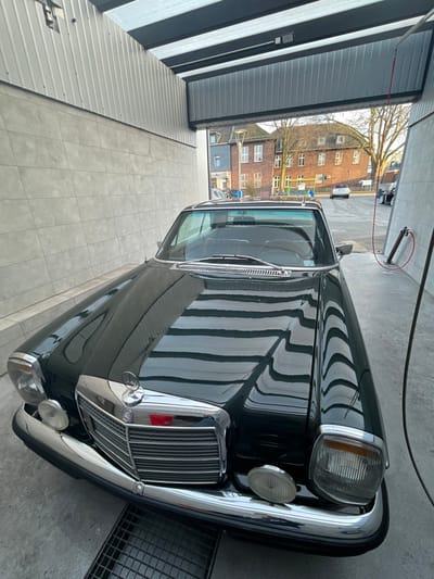 A dark green vintage Mercedes-Benz, likely a W111 or W108/109 series, is parked front-first within a covered wash bay in Oberhausen, Germany. The vehicle's polished hood prominently reflects horizontal striped shadows from the overhead structure. The bay features light gray tiled walls, a concrete floor with a metal drainage grate, and industrial corrugated metal paneling on the roof. On the right, a red hose hangs from an overhead dispenser towards a vertical metal wash station fixture. Beyond the bay, an urban street is visible under natural light, lined with traditional multi-story brick buildings, bare trees, and other parked cars. No individuals are present.