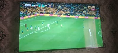 The image shows a television screen displaying a soccer match. The score is UZB 1, COL 2, with the time at 87:28. There are players in green and white uniforms on the field, and a referee in a yellow shirt. The stadium appears to be full of spectators, many wearing yellow. The ZOR TV logo is visible in the upper right corner, along with the word "LIVE." The scene is set in a soccer stadium. The city of Kasan, Uzbekistan is not depicted in this image.