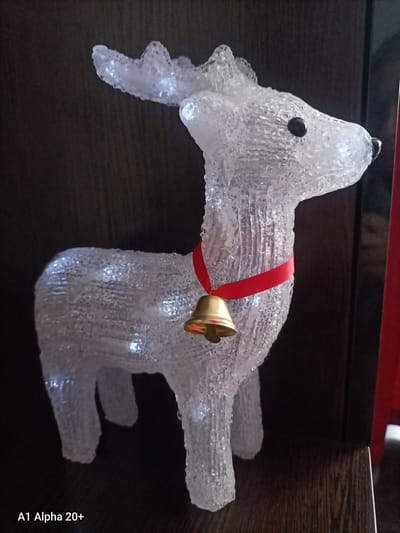 An illuminated, clear plastic reindeer decoration with white lights is positioned on a dark wooden surface. The reindeer has a red ribbon around its neck with a small golden bell hanging from it. The background is a dark, textured surface, likely wood paneling. The lighting suggests an indoor setting. The image is a still photograph, and there is no visible activity or interaction occurring. There are no people present in the image. The image was captured in Улукаўскі сельскі Савет, Belarus.