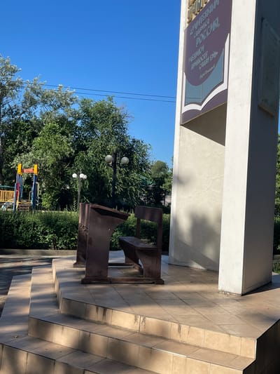 In Pokoinoe, Russia, a memorial composed of a large, off-white concrete structure stands under a clear blue sky. A plaque with text in Cyrillic script, featuring a graphic of an open book, is affixed to the monument. In front of the monument, a weathered, dark metal bench and desk unit sits on a tiled platform with several steps. To the left, a colorful children's playground structure is visible amidst lush green trees and bushes. The scene is illuminated by bright sunlight, casting distinct shadows.