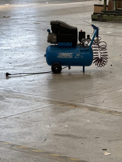 A blue air compressor with a black motor unit sits on a concrete floor in an industrial or warehouse setting. The compressor has a visible "ABAC 24 Hp 2" label. A black power cord extends from the unit to the left, and a coiled dark red hose is attached to the compressor, leading to a blue blow gun. The concrete floor appears worn, with visible lines and some debris scattered around. In the background, there are shelves and boxes, suggesting a storage area. The overall lighting suggests an indoor environment, possibly a workshop or a storage facility in Fontevivo, Italy. There are no people visible in the image.