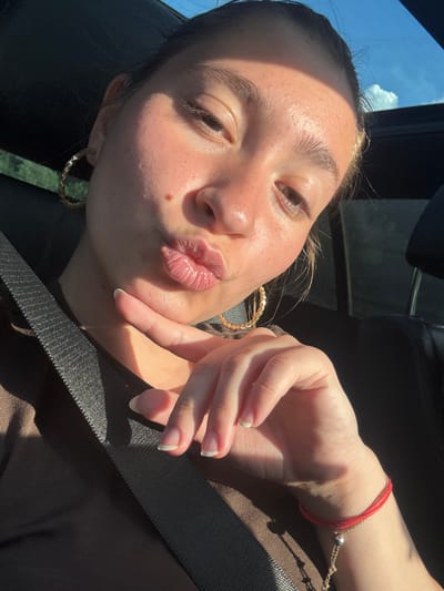 The image is a close-up, indoor, selfie-style photograph of a young woman with her hand on her chin, puckering her lips. She is wearing large hoop earrings and a red string bracelet on her wrist. A seatbelt is visible across her chest. The background suggests she is in a car, with a dark interior and a glimpse of a blue sky with clouds through the sunroof. The lighting indicates it is daytime. There is no indication of the location being the city of Dolno Varshilo, Bulgaria.