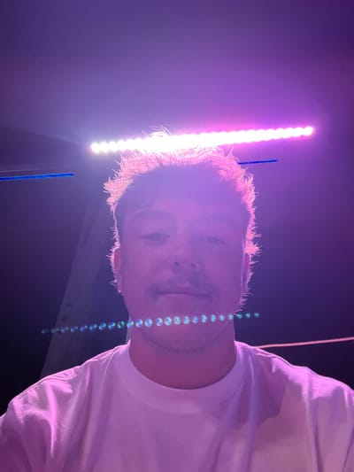A single male individual, appearing to be in their 20s or 30s, is depicted in a front-facing portrait, looking directly at the camera. The subject has light-colored, medium-length hair, illuminated by a strong backlight, and a visible mustache. They are wearing a plain white crew-neck t-shirt. The scene is dark, indicating a nighttime or low-light environment. Above the subject's head, a prominent horizontal light fixture, composed of individual LED lights, emits bright pink, purple, and white illumination, which backlights the subject's hair and casts a magenta glow over the image. Optical artifacts are present, including a faint horizontal blue line in the upper-left portion of the image and a distinct horizontal string of circular, iridescent lens flares appearing below the subject's chin. A vertical structural element is partially visible on the left side of the background, and a faint horizontal line is visible on the right. This scene is located in Albufeira, Portugal.