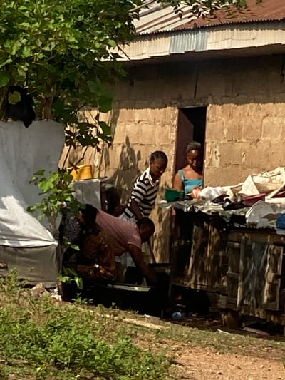 The image captures an outdoor scene in Idiroko, Nigeria, featuring a single-story building and four individuals engaged in activities. The building, constructed of light-colored concrete blocks, has a corrugated metal roof and a dark, open doorway.
In the background, a female wearing a light blue top is partially visible within the doorway, looking downwards. In front of the doorway, a young person in a black and white striped polo shirt stands, looking down and to the left. In the mid-ground, a female wearing a pink shirt and a patterned wrapper is bent forward, tending to a cooking setup on the ground. This setup includes a dark pot positioned over a visible flame, with a blue bottle nearby. A fourth individual is partially obscured behind the bending female.
To the right of the people, a makeshift counter or table holds various fabrics, including white and blue pieces, and a green bowl. The left side of the frame is dominated by dense green foliage, partially obscuring a white tarp-like structure and a yellow bucket. The ground consists of dirt with patches of green vegetation. Bright sunlight illuminates the scene, casting shadows from the foliage onto the building.