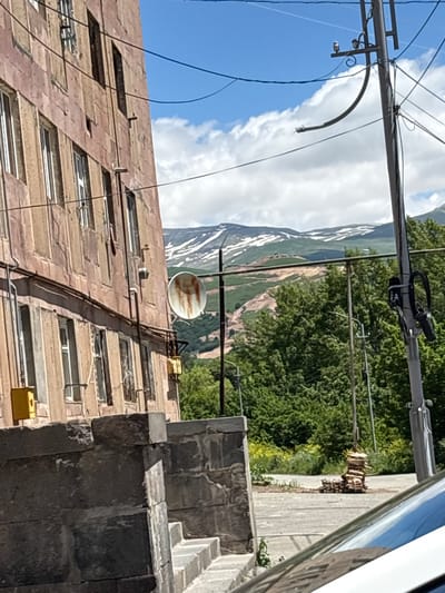 In a medium-shot, outdoor scene taken on a bright, partially cloudy day in Jermuk, Armenia, a weathered, pinkish-brown apartment building occupies the left side of the frame, its facade marked by numerous windows with white frames and metal bars. A rusty, circular satellite dish is mounted on the building's exterior, to the right of which is a yellow electrical box. In the foreground, a section of a stone wall and steps is visible. Beyond the building, a chain-link fence runs horizontally across the middle ground. In the distance, lush green trees line the base of a mountain range, the upper slopes of which are partially covered in snow. A utility pole with wires and attached equipment stands prominently in the right middle ground, next to a stack of firewood. The overall environment suggests an urban setting with a natural backdrop. There are no people visible in the image.
