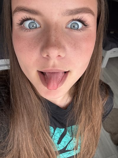A close-up, head-on shot captures a young person with light brown hair, fair skin, and freckles, their eyes wide and blue, looking directly at the camera. Their tongue is extended. They are wearing a dark t-shirt with a teal geometric pattern. The background is blurred, showing hints of a room with light-colored walls and furniture. No specific location in Shepetivka, Ukraine is discernible.